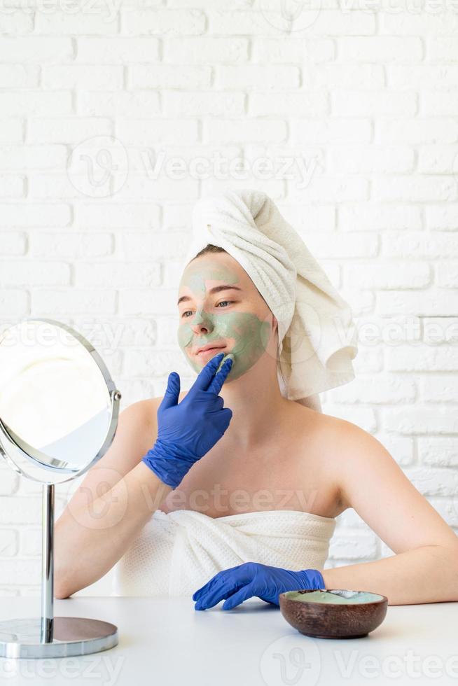 Woman wearing gloves applying clay face mask looking at the mirror photo