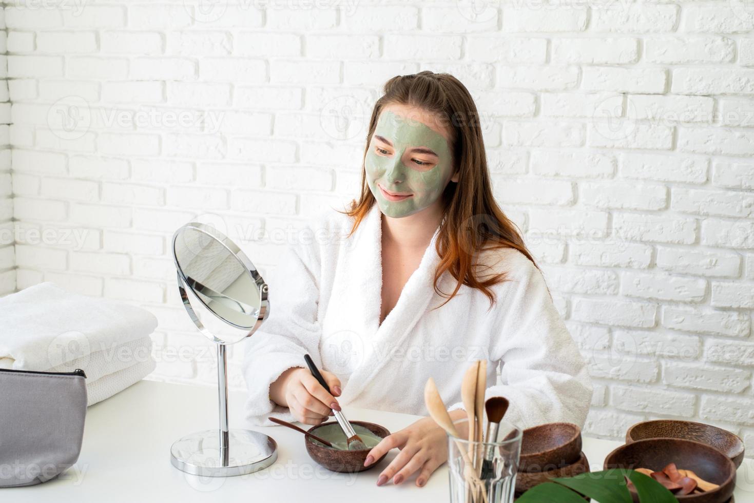 Mujer vistiendo albornoces aplicando mascarilla de arcilla mirando el espejo foto