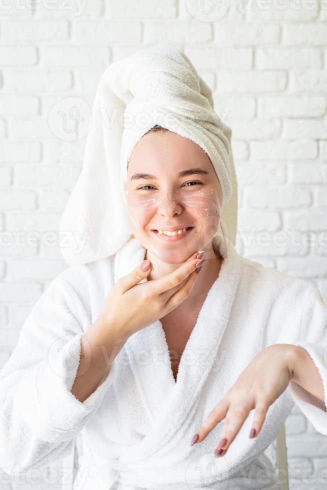 Caucasian woman in white bath towel applying face cream at home photo