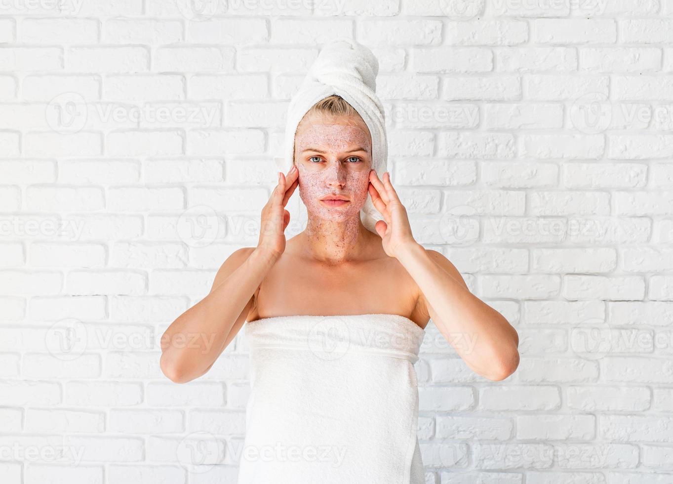 Mujer joven en toallas de baño blancas aplicando exfoliante en la cara y el cuello foto