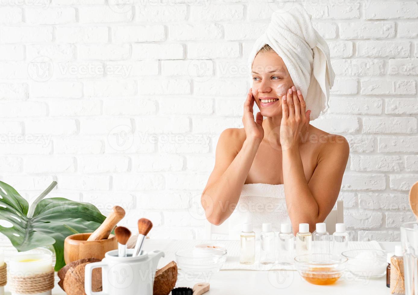 Mujer en toalla de baño blanca aplicando crema facial haciendo procedimientos de spa foto