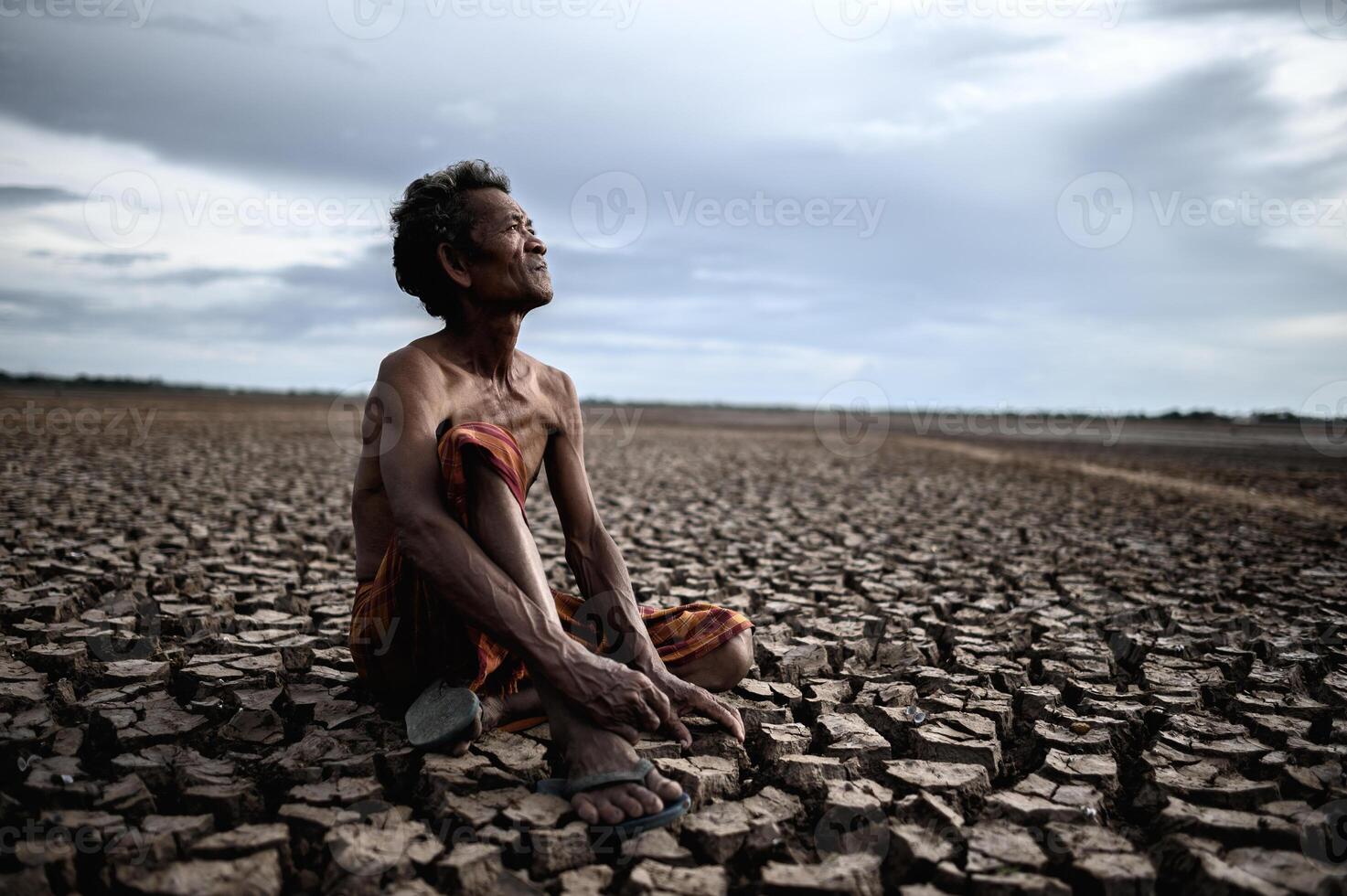 An elderly man sat hugging his knees bent on dry soil photo