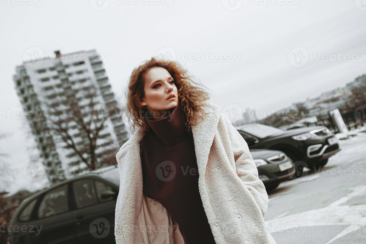 A girl with red curly hair in white coat poses on outdoor parking photo