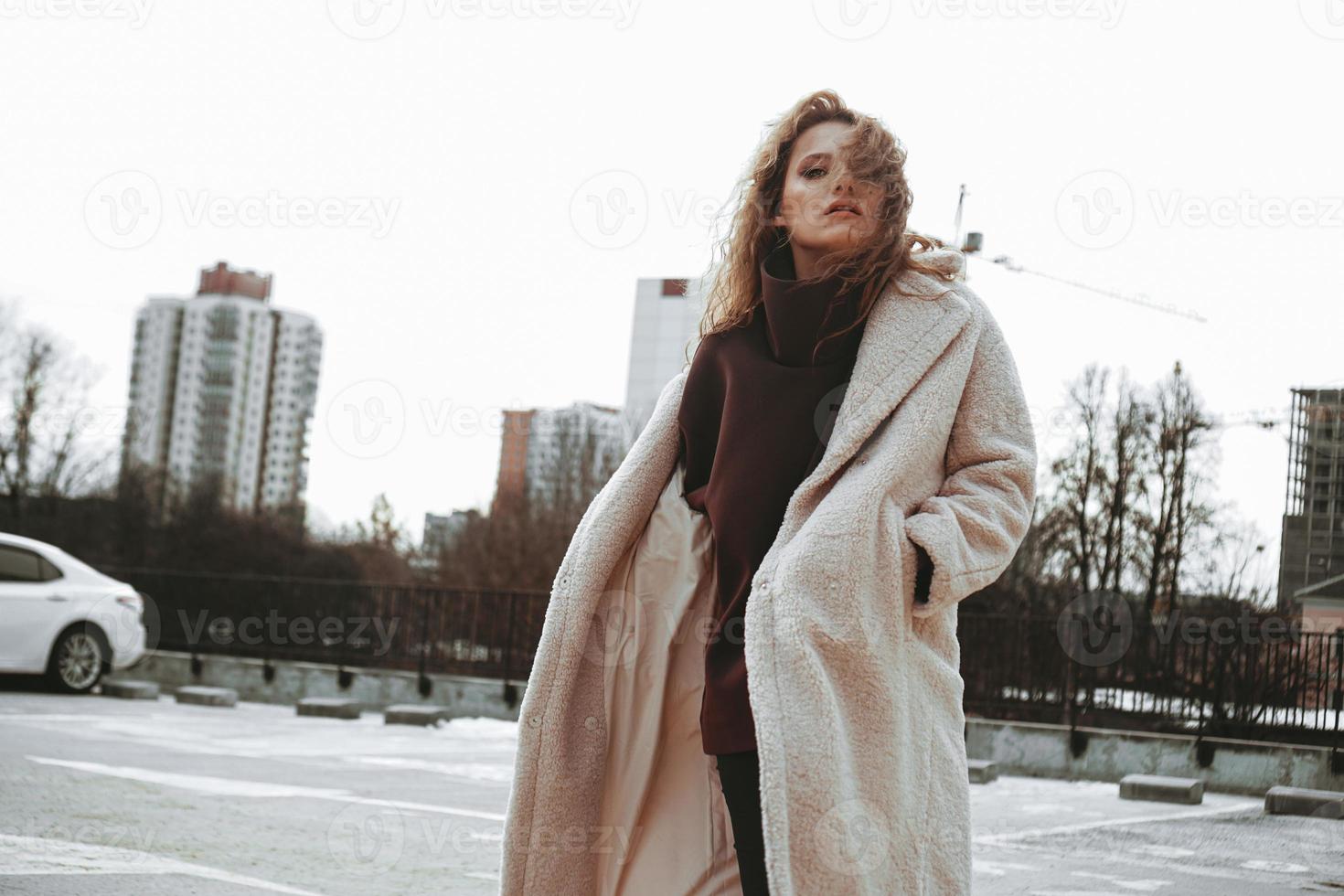 A girl with red curly hair in white coat poses on outdoor parking photo