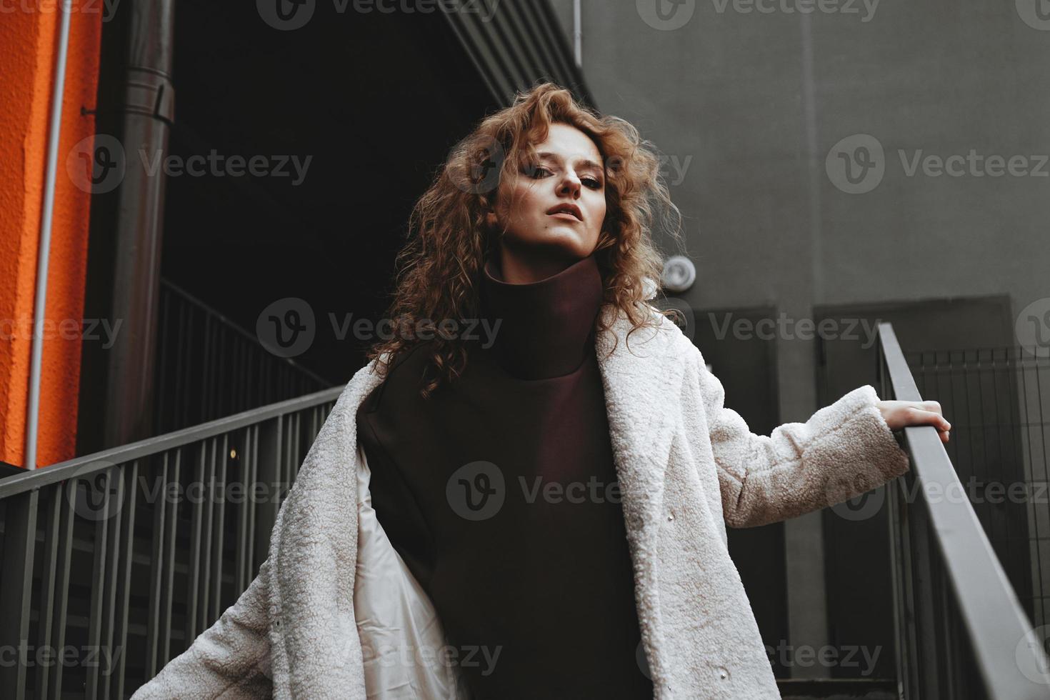 una chica con el pelo rojo y rizado con una bata blanca posa en las escaleras del estacionamiento foto