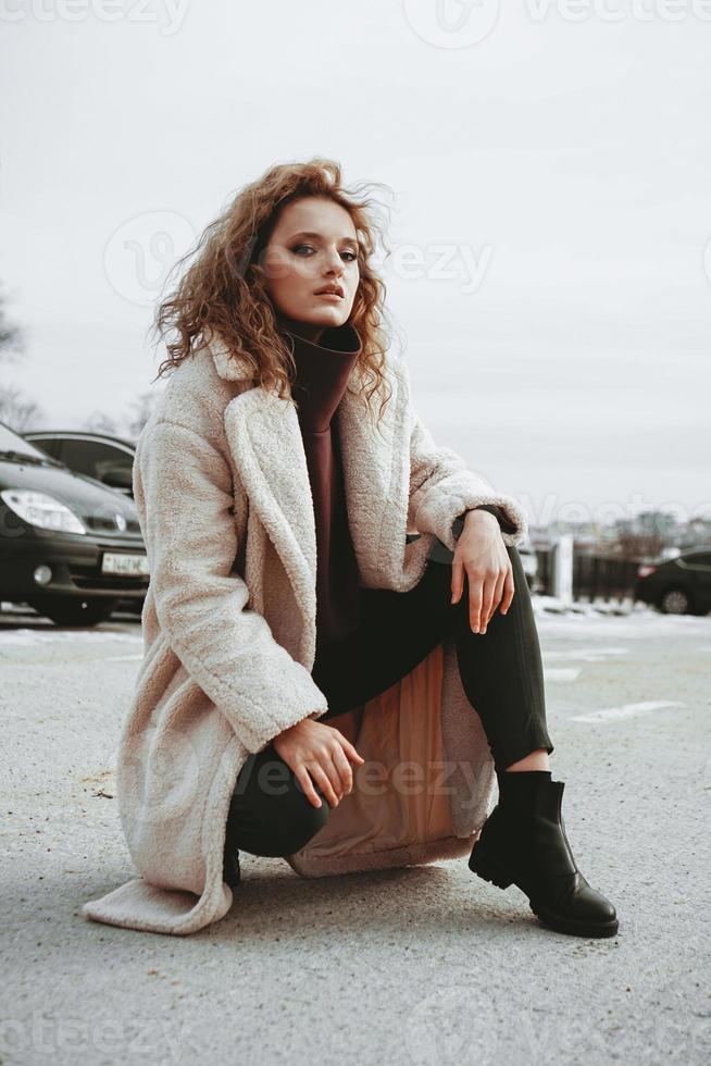 A girl with red curly hair in white coat poses on outdoor parking photo