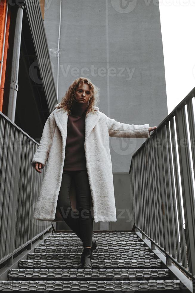 una chica con el pelo rojo y rizado con una bata blanca posa en las escaleras del estacionamiento foto