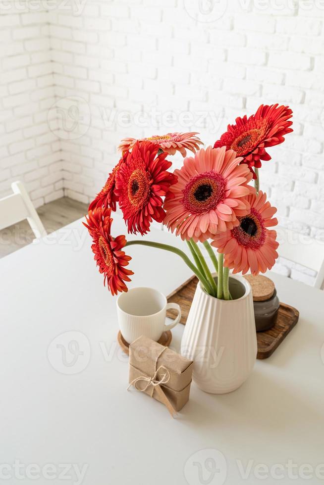 Bright gerbera daisies in white vase on kitchen table with a gift box photo