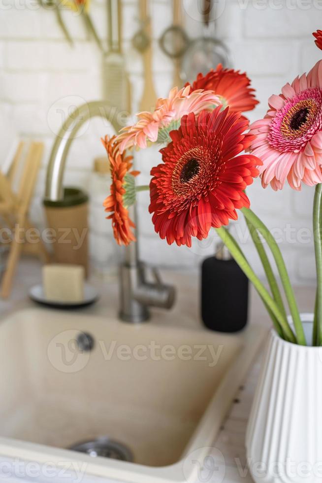 Red and pink gerbera daisies in a white vase on a wooden kitchen photo