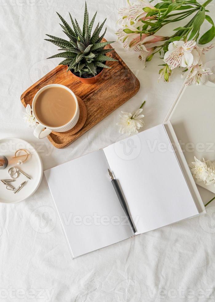 Opened book, coffee and flowers top view on white bed. Mock up design photo