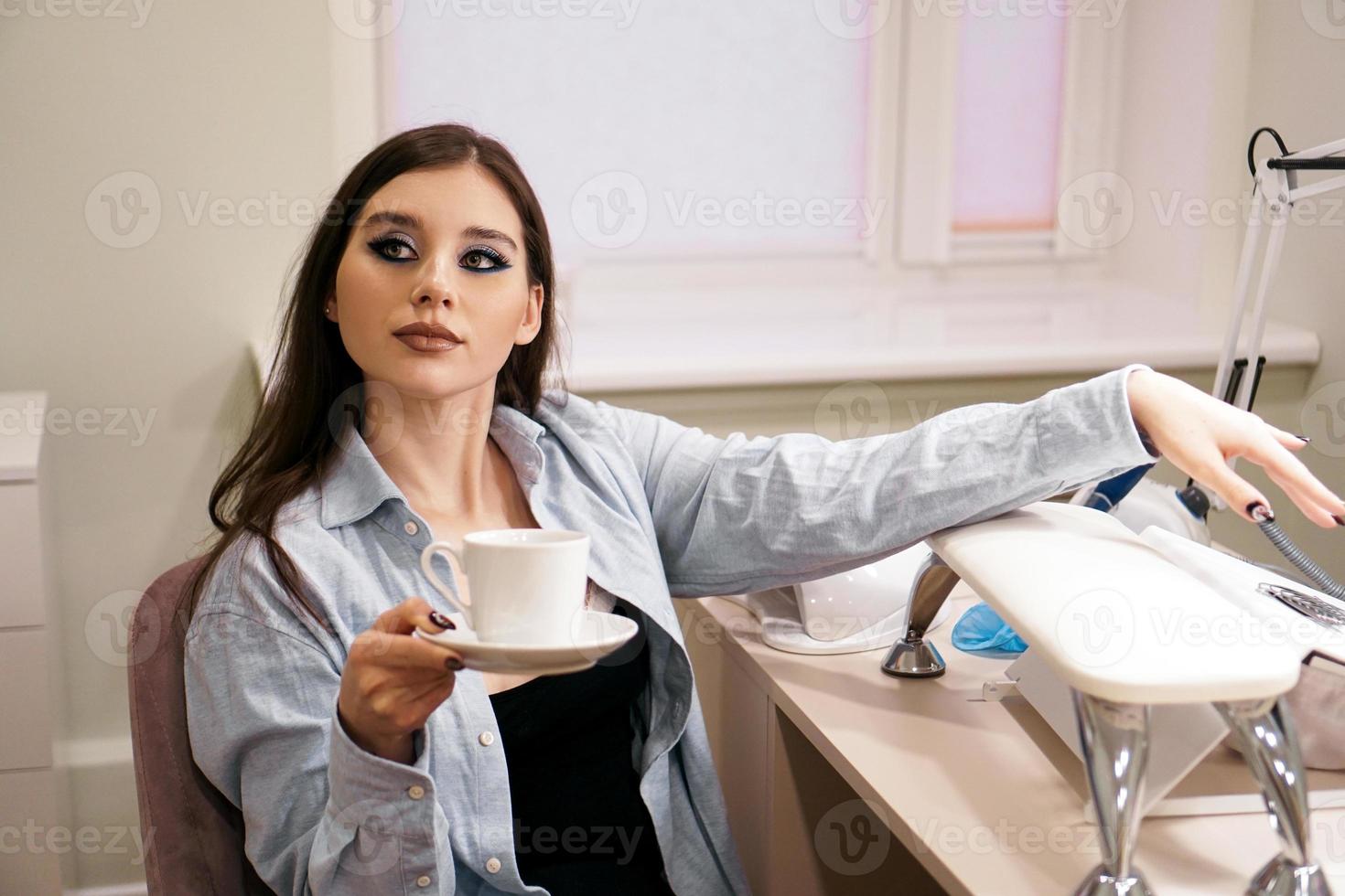 Woman does manicure in a beauty salon and holds a white cup of coffee photo