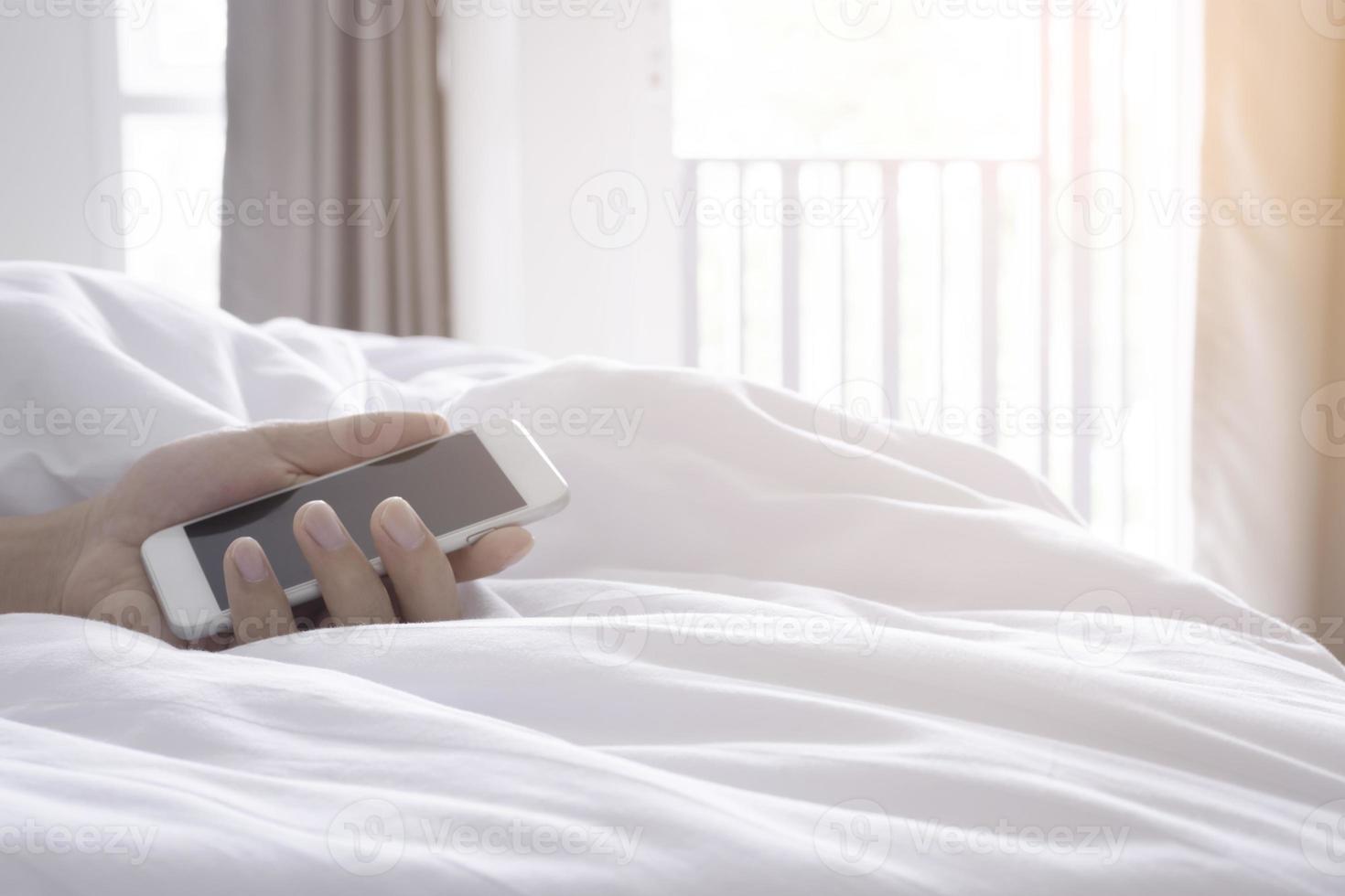 mano que sostiene el teléfono inteligente en la cama blanca en la mañana foto