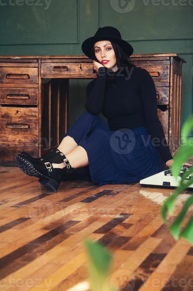 Hermosa mujer con sombrero se sienta cerca de la mesa de roble con máquina de escribir vintage foto