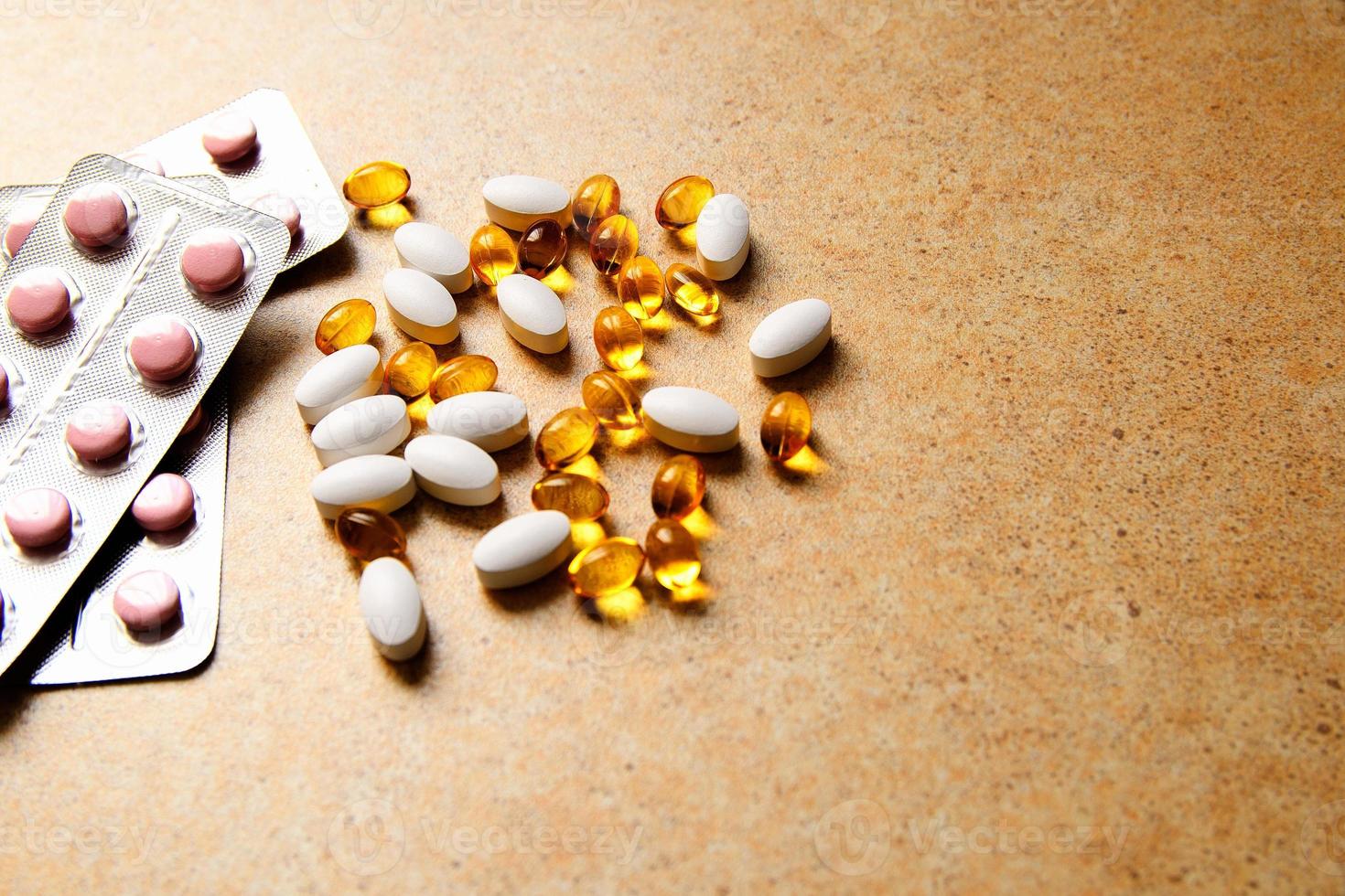 Blister with pink tablets and vitamin D and capsules with fish oil lie chaotically against the background of a sand countertop photo