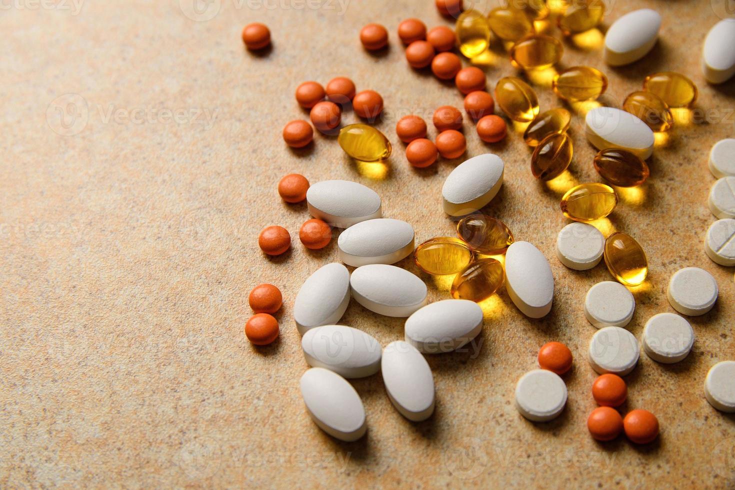 Orange pills, vitamin D and fish oil capsules lie randomly against a sand countertop photo