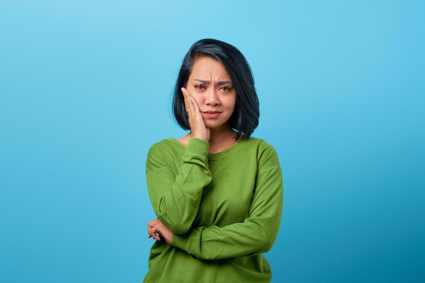 Mujer asiática tocando la mejilla con expresión aburrida sobre fondo azul. foto
