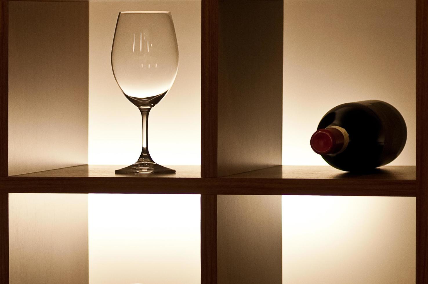 A single empty wine glass with beautiful reflections and a closed bottle of red wine lying on a shelf with side lighting set in the interior photo