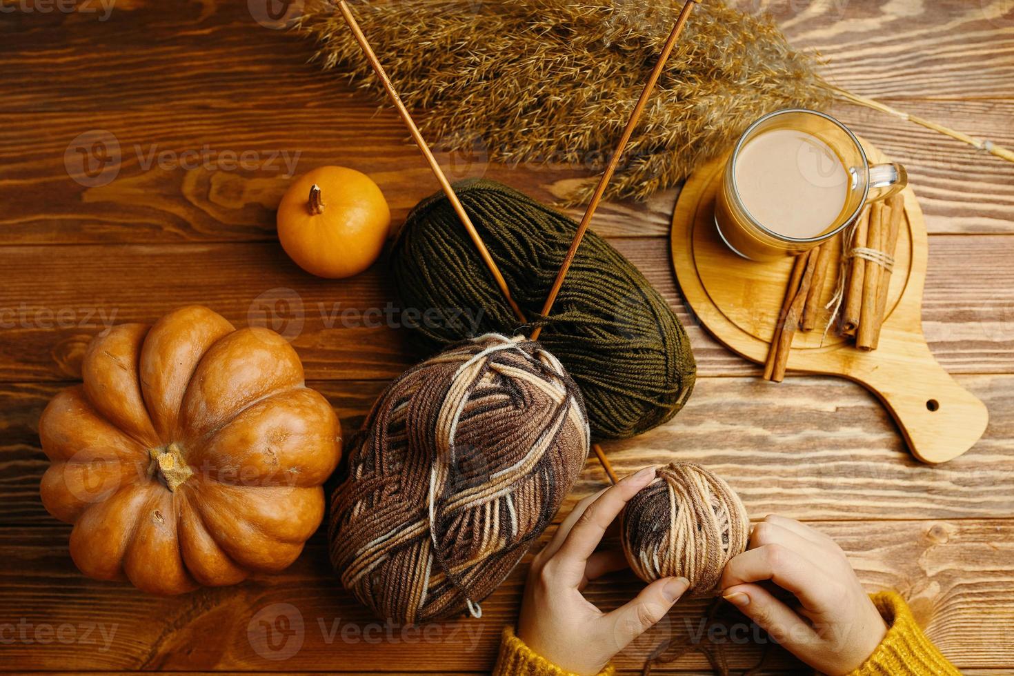 Manos en suéter naranja con hilo, agujas de tejer, café y palitos de canela en la mesa de madera foto