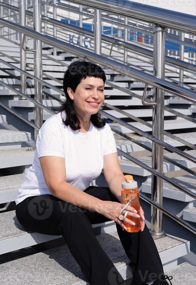 Senior woman drinking water after workout outdoors on urban background photo