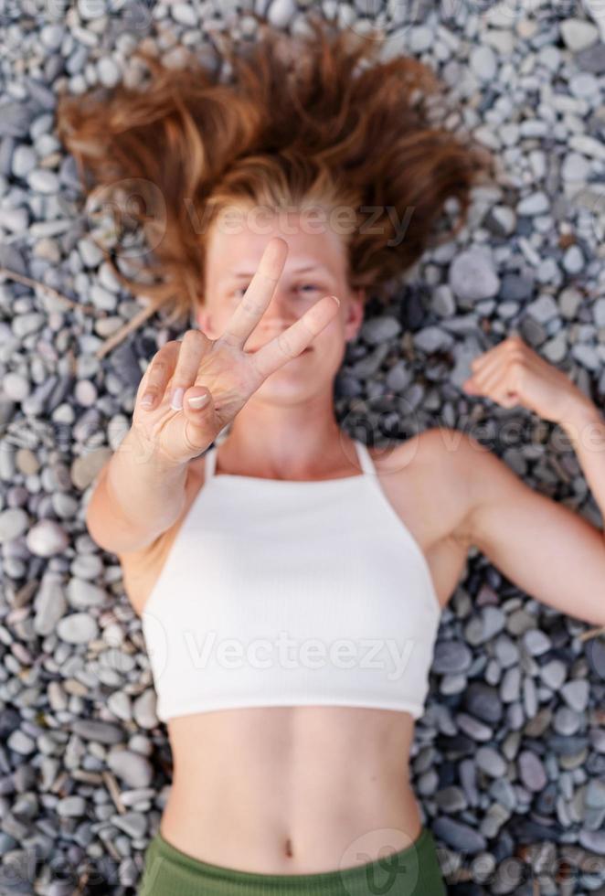 Mujer sonriente joven tumbado en la playa de piedra, cubriendo el rostro con las manos foto