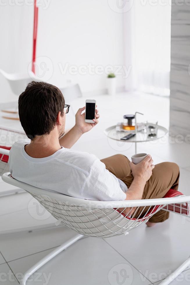 Man sitting in a chair watching tv holding tea cup and phone photo