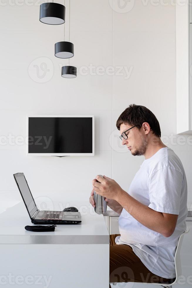 Young smiling man working with laptop from home, drinking water photo