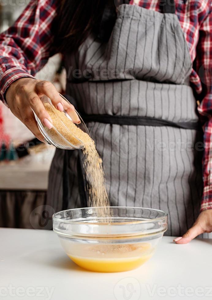 Joven mujer latina vertiendo azúcar en la masa para cocinar en la cocina foto