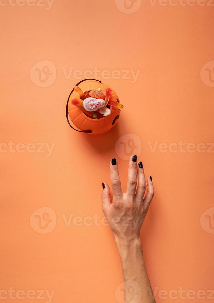 Scary woman's hand with black nails trying to get pumpkin with sweets, flat lay on orange background photo