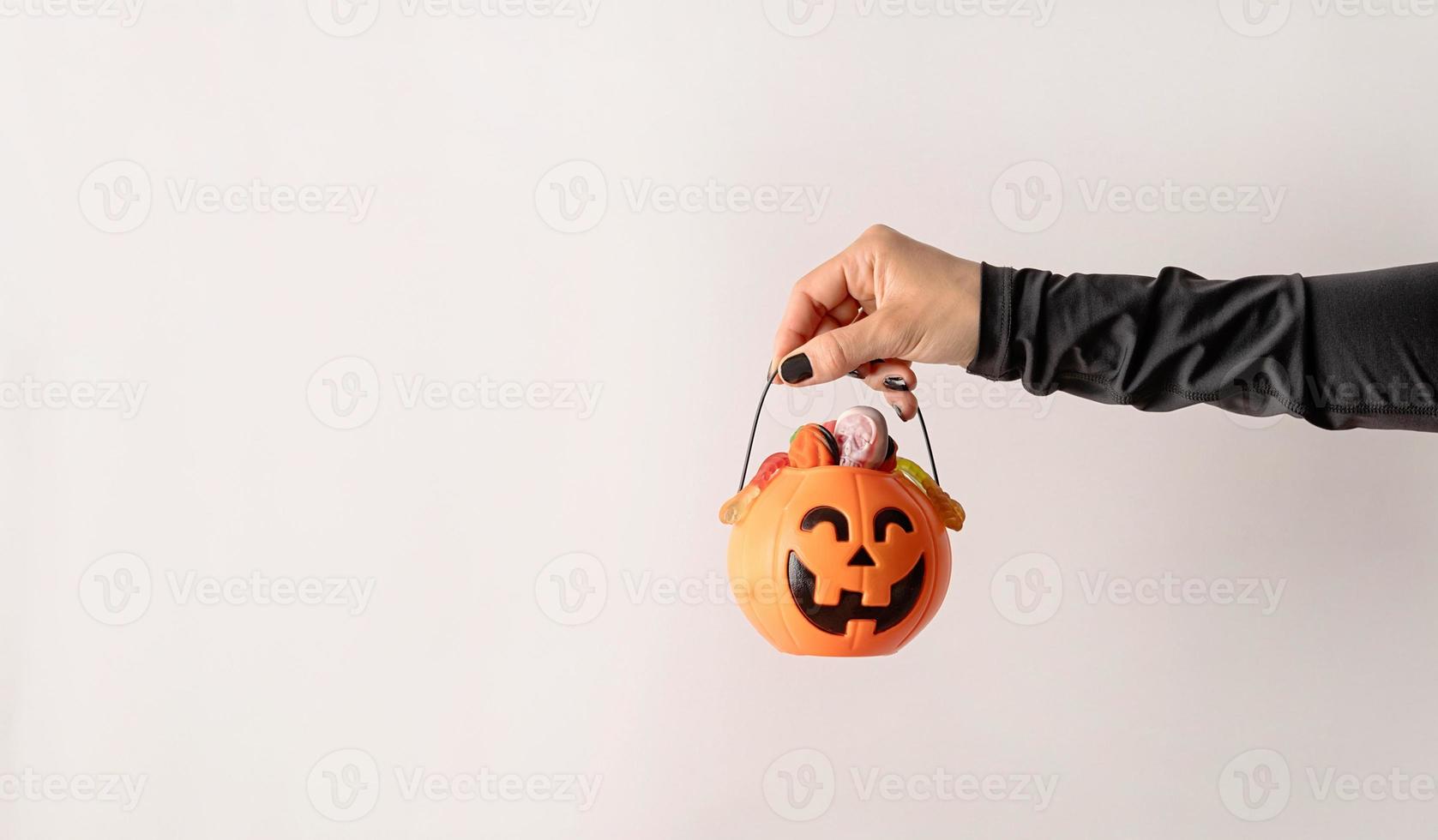 Mano de mujer con uñas negras sosteniendo calabaza llena de dulces sobre fondo blanco. foto