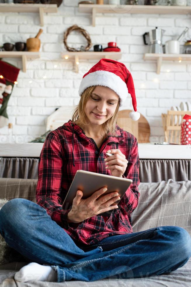 Joven mujer rubia con gorro de Papá Noel trabajando en tableta sentado en el sofá foto