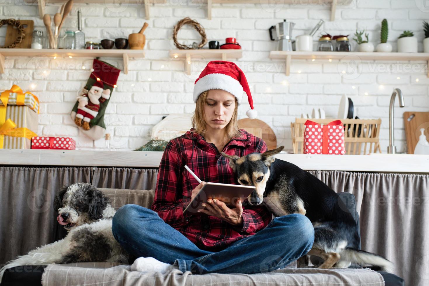 Joven mujer rubia con gorro de Papá Noel trabajando en tableta sentado en el sofá foto