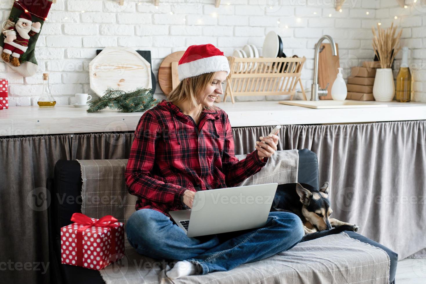 Young blonde woman in santa hat working on laptop looking at phone photo