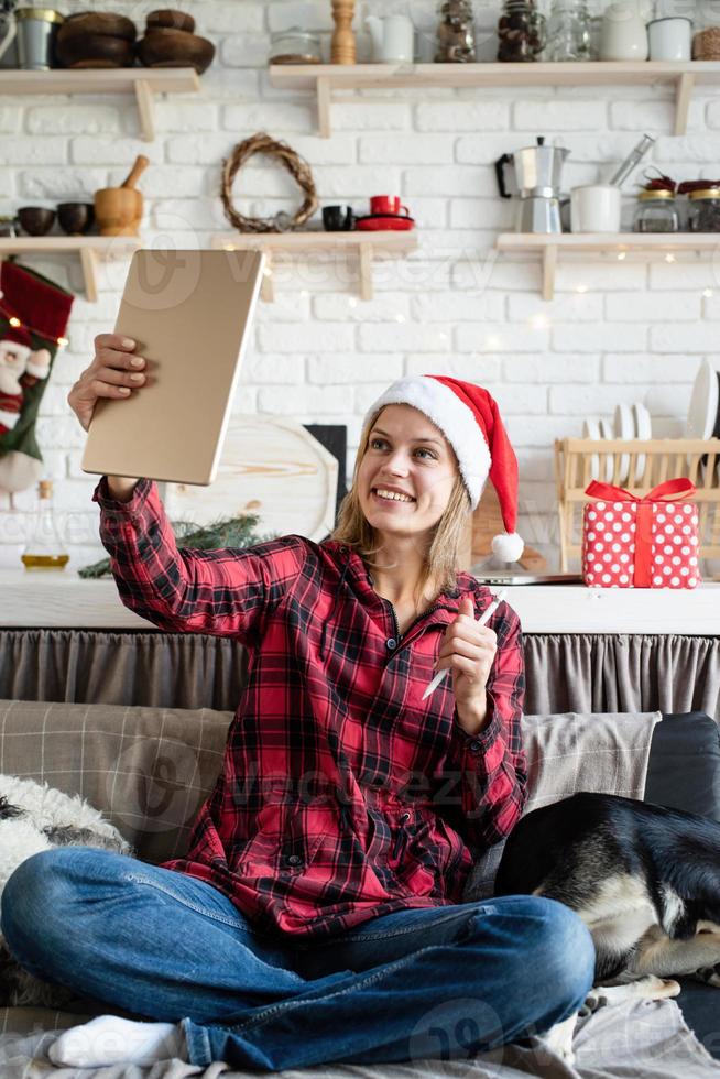 Joven mujer rubia con gorro de Papá Noel trabajando en tableta sentado en el sofá foto