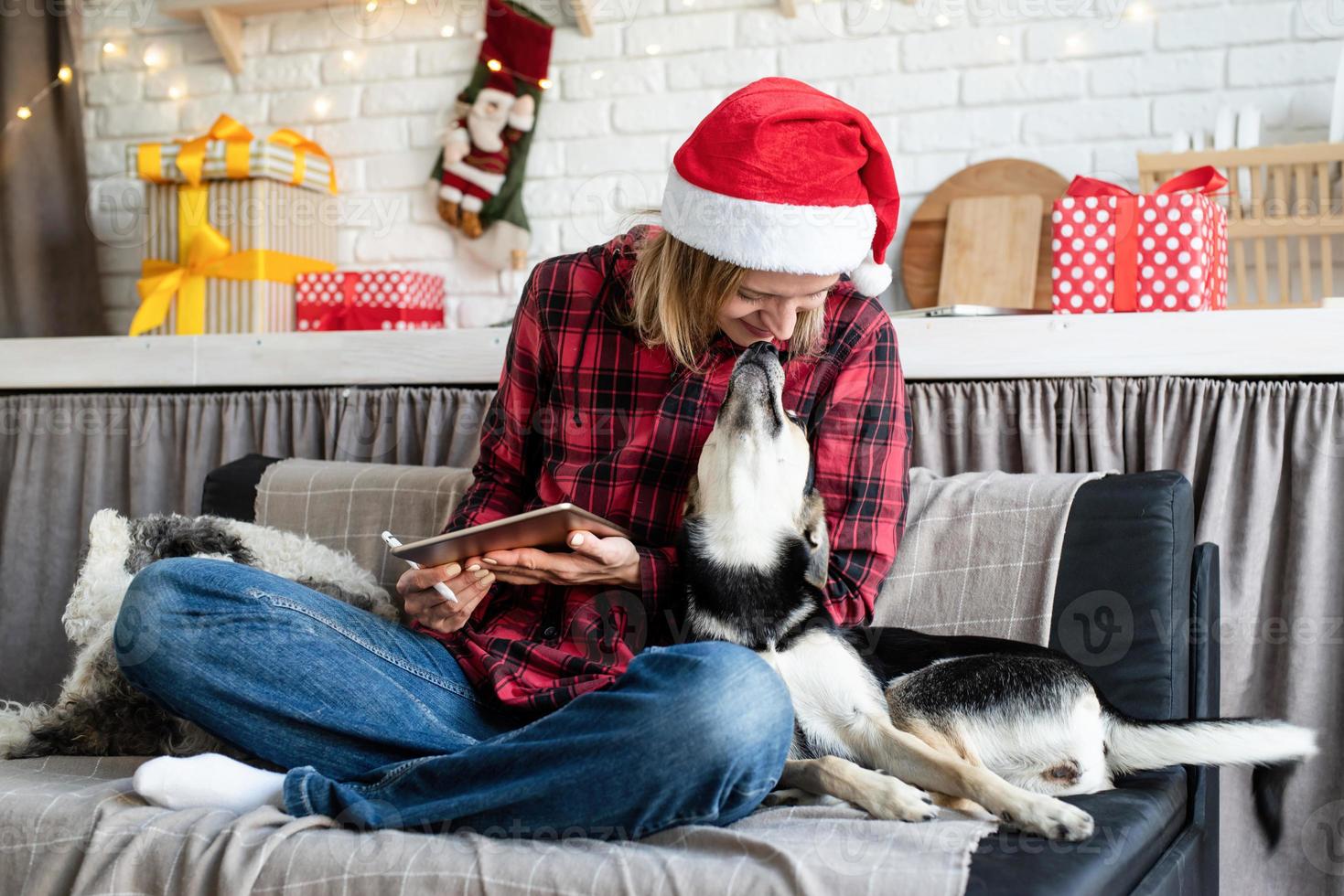 Mujer con gorro de Papá Noel trabajando en tableta sentada en el sofá con su perro foto