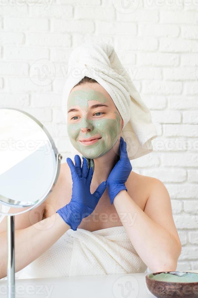 Mujer con guantes aplicando mascarilla de arcilla mirando el espejo foto