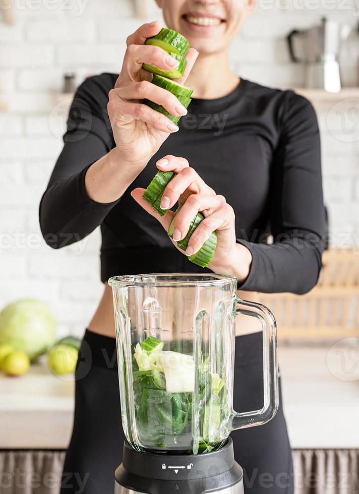 Young blond smiling woman making cucumber smoothie at home kitchen photo