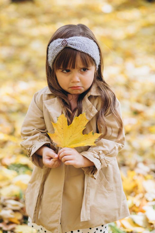 niña ofendida sosteniendo una hoja de arce amarilla en el parque de otoño foto