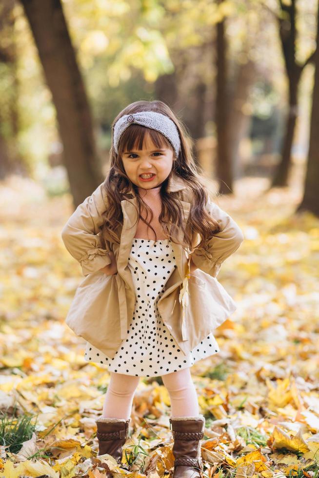 Beautiful little girl in a beige coat shows angry in the autumn park photo