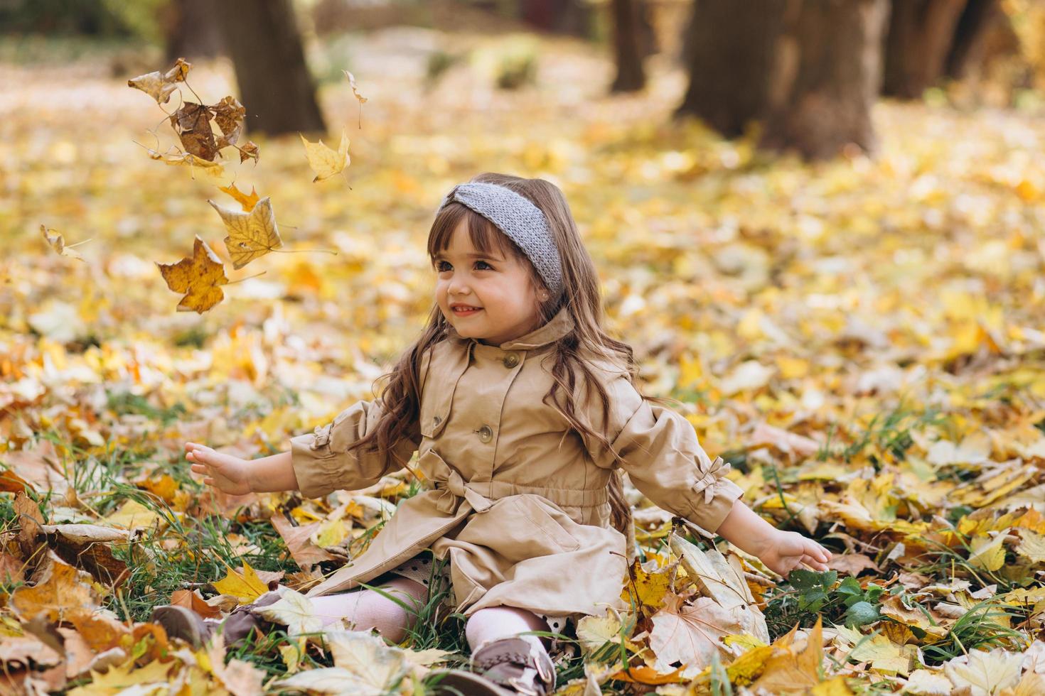 niña con un abrigo beige sentada entre hojas en el parque de otoño. foto