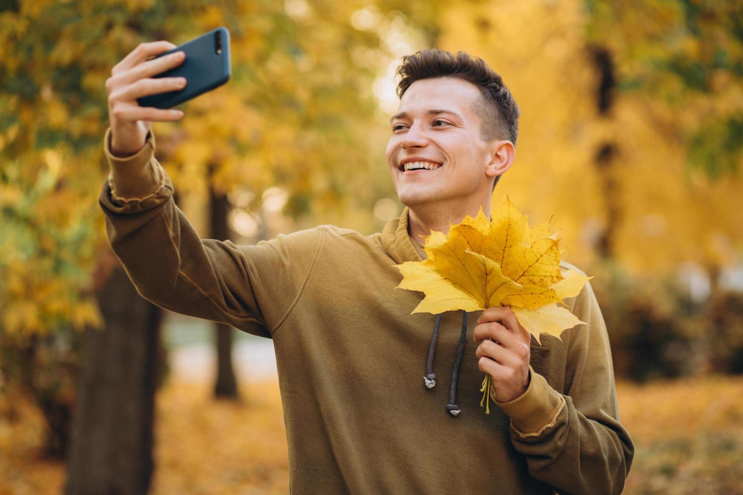 Chico sonriendo y sosteniendo un ramo de hojas y tomando selfie en el parque foto