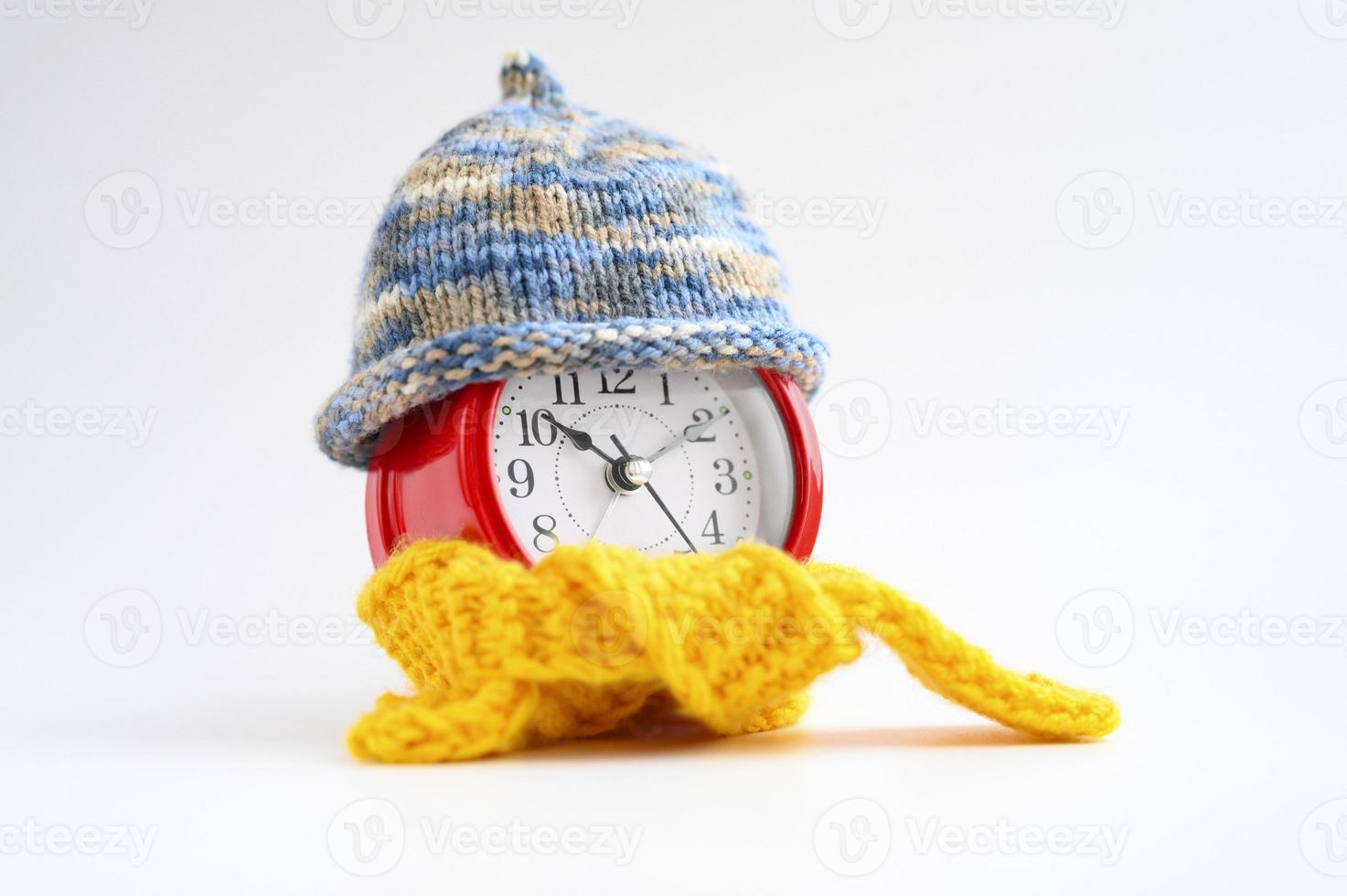 Red round alarm clock in knitted wool blue hat and yellow scarf on a white background. wintertime concept. winter season. cozy and warm. analog time 10 10. space for text. banner photo