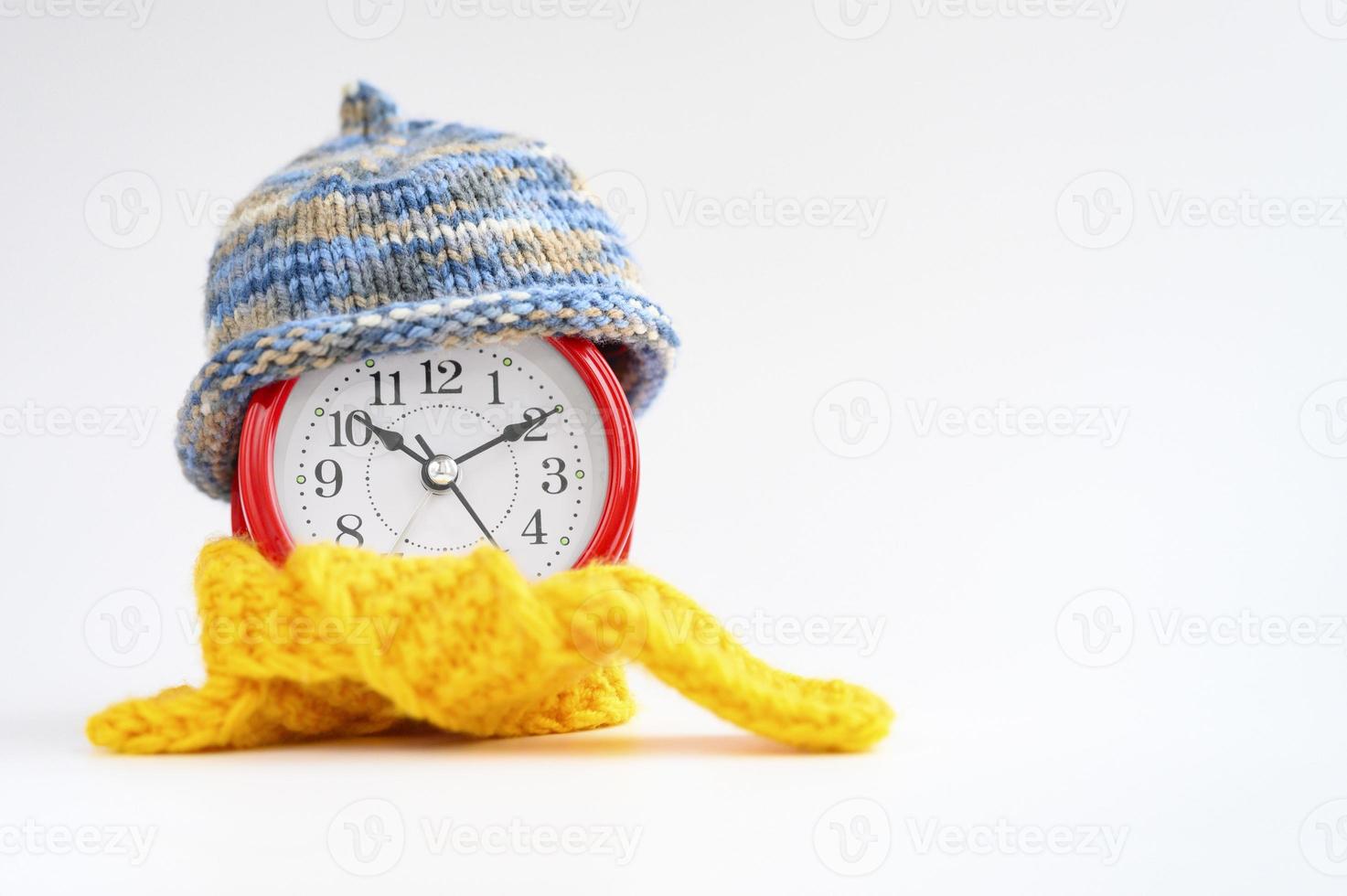 Red round alarm clock in knitted wool blue hat and yellow scarf on a blue background. wintertime concept. winter season. cozy and warm. analog time 10 10. space for text. banner photo