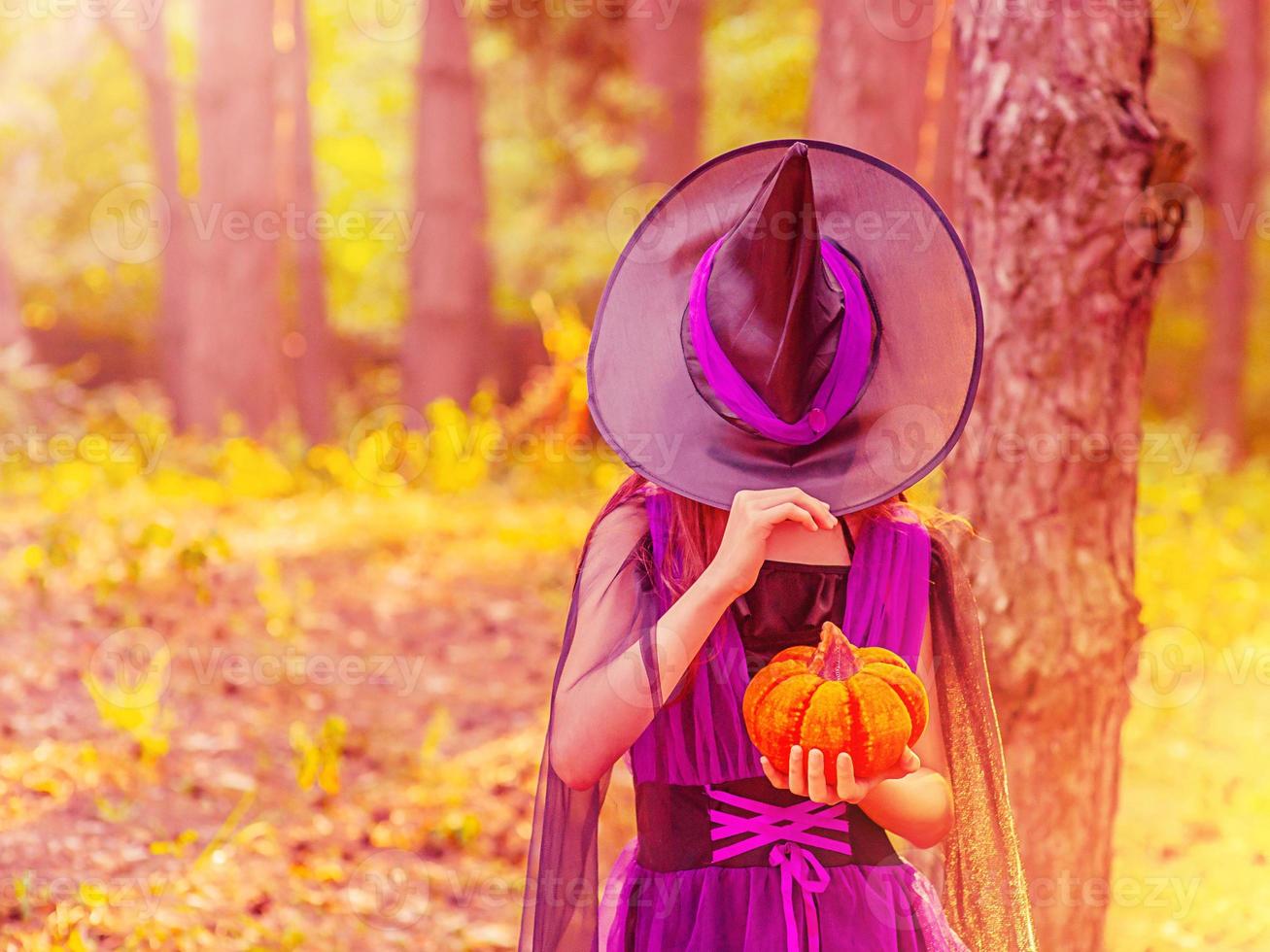 niña en disfraz de halloween con sombrero sobre los ojos con calabaza naranja foto