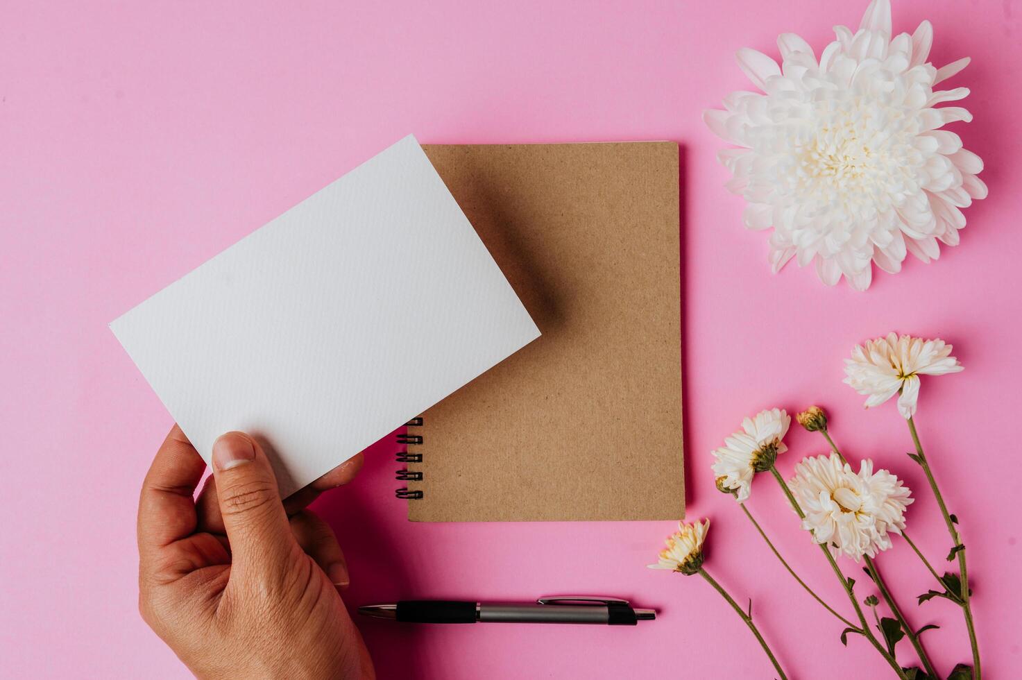 mano que sostiene la tarjeta en blanco, cuaderno, bolígrafo y flor sobre fondo rosa foto