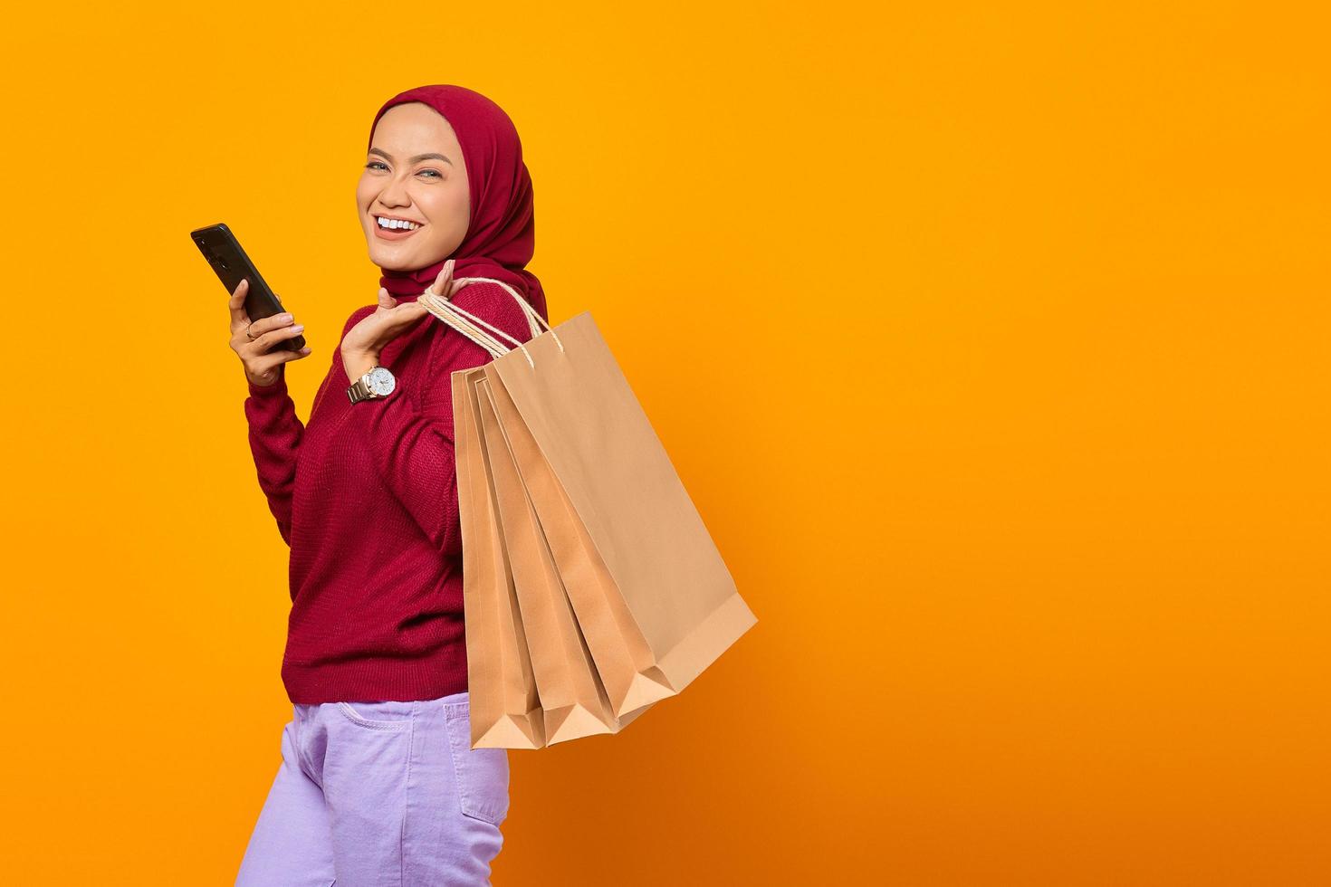 Cheerful asian woman holding smartphone and showing shopping bags photo