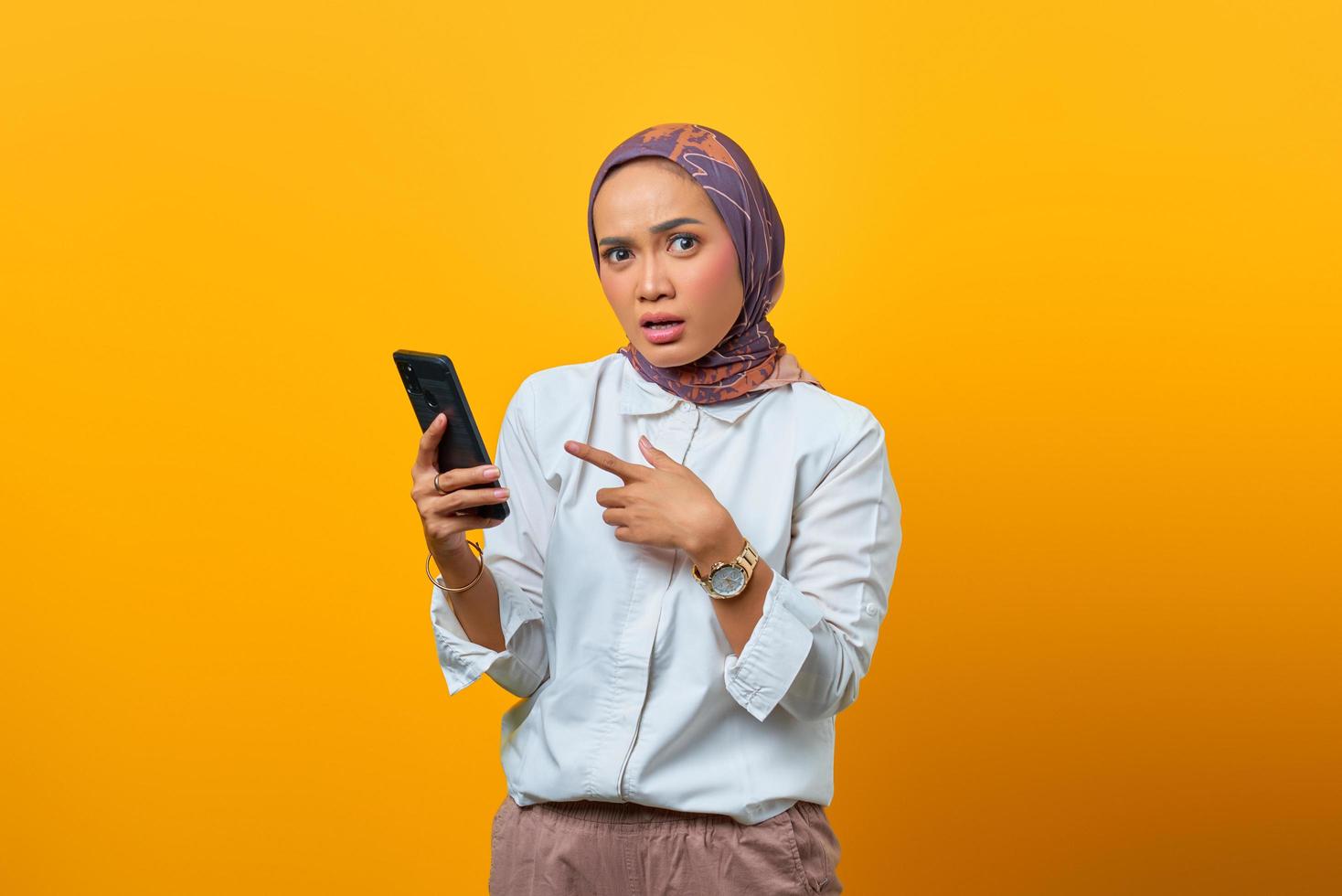 Surprised Asian woman while pointing smartphone over yellow background photo