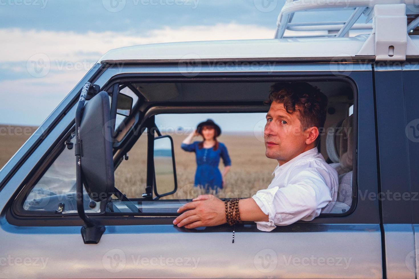 Hombre conduciendo un coche y una mujer con vestido y sombrero caminando por el campo de trigo foto