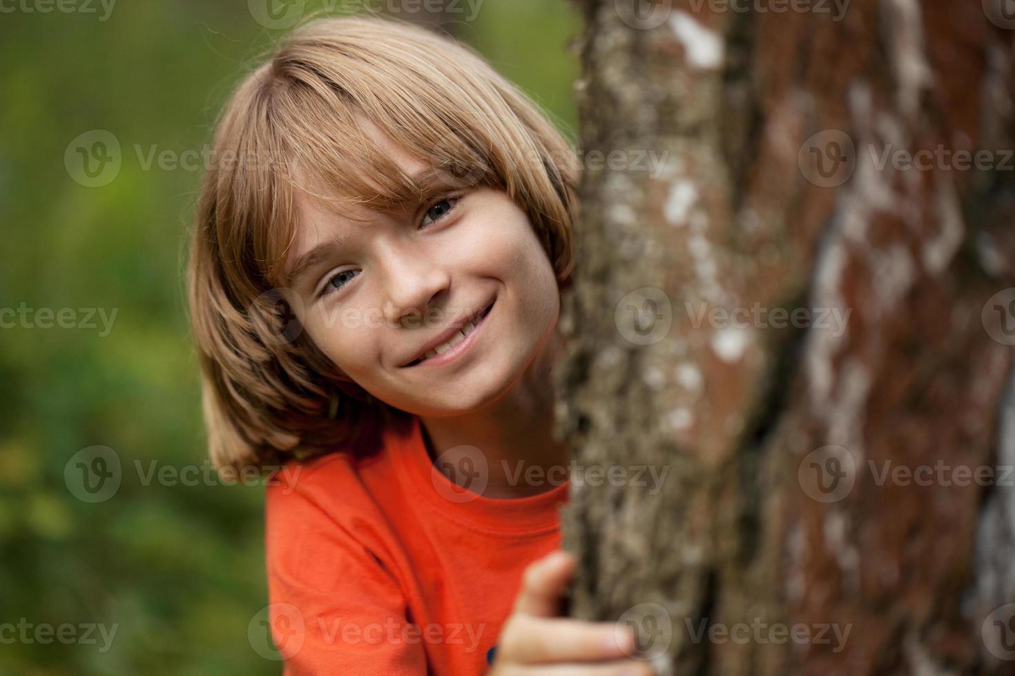 Boy in red T-shirt peeking out from behind a tree trunk photo