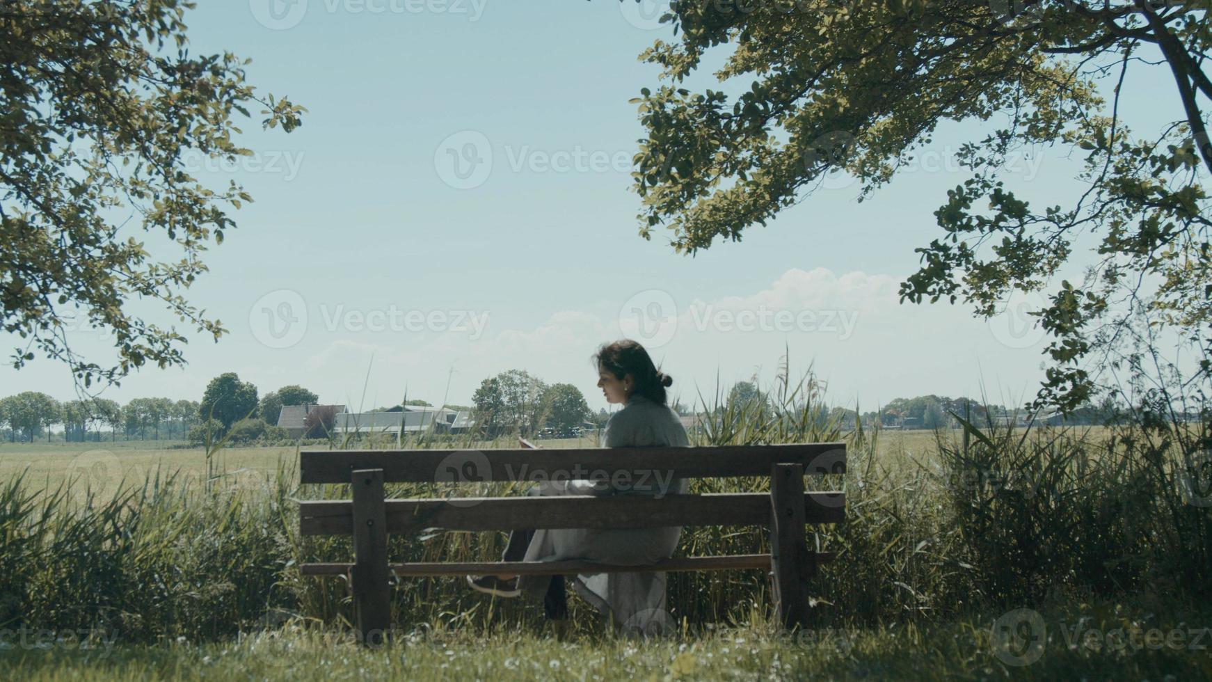Woman sits down on wooden bench in the country and starts reading book photo