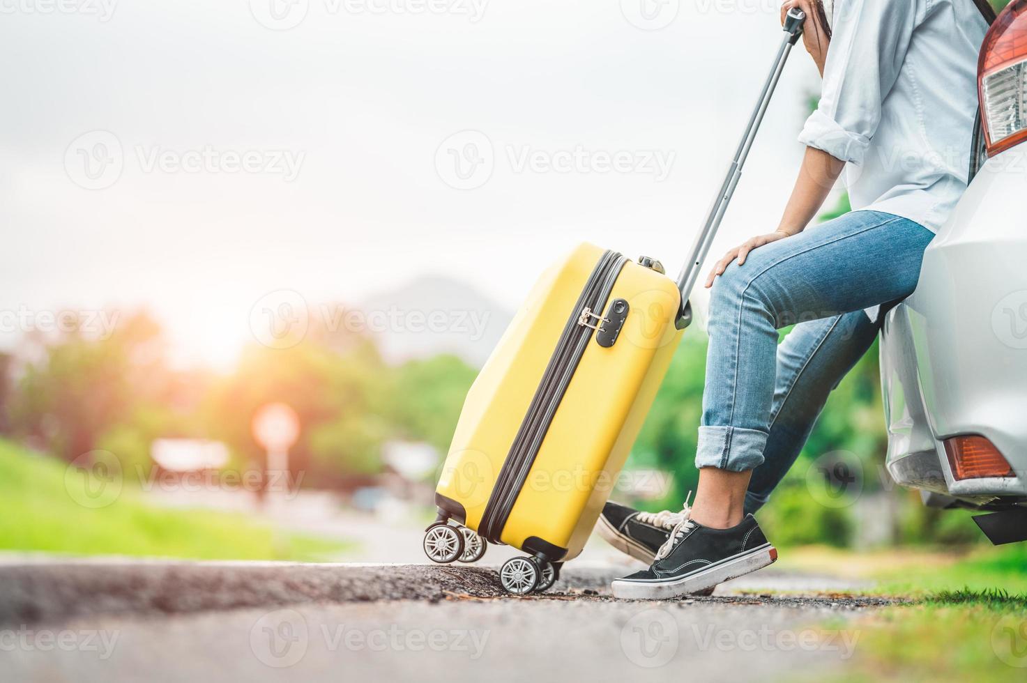 Primer plano de la parte inferior del cuerpo de la mujer relajándose en el maletero del coche con el carrito de equipaje foto